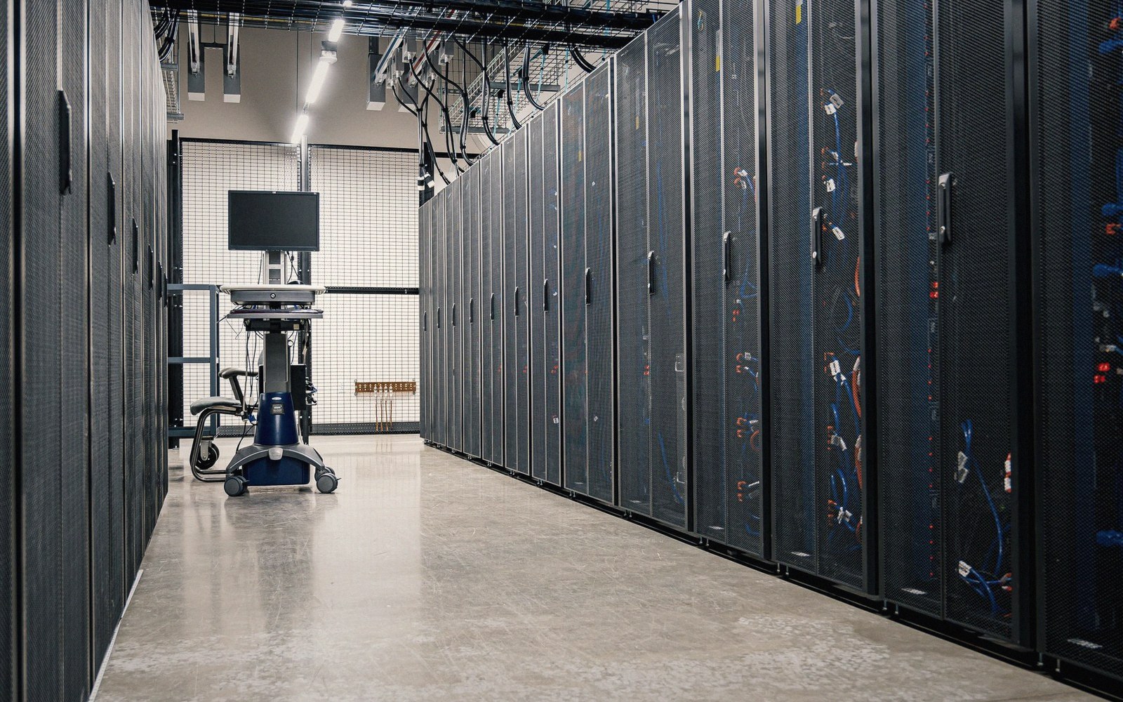 A server rack aisle with dense cable management in blues and yellows, shot from floor level looking toward a hot-aisle containment door